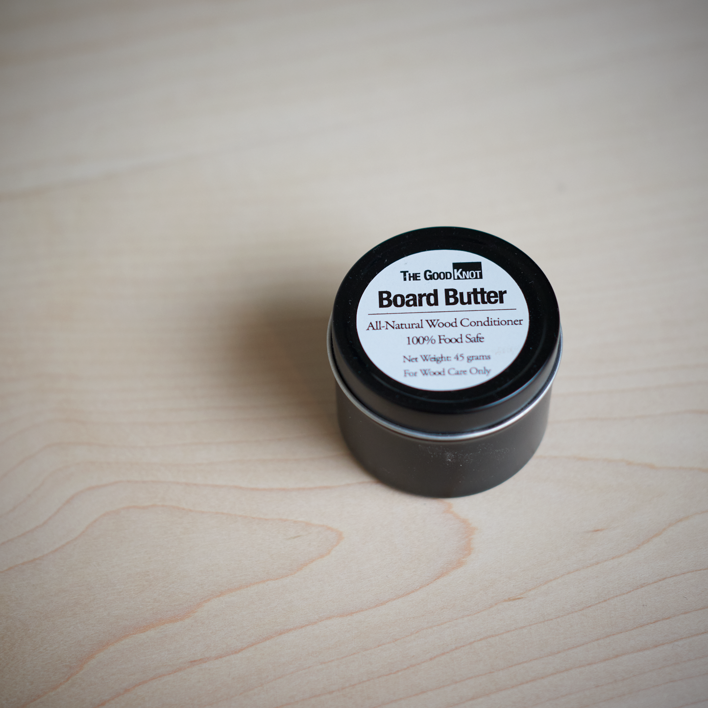 A container of all-natural wood conditioner, Board Butter, with a label that reads 'The Good Knot', is shown on a wooden surface.