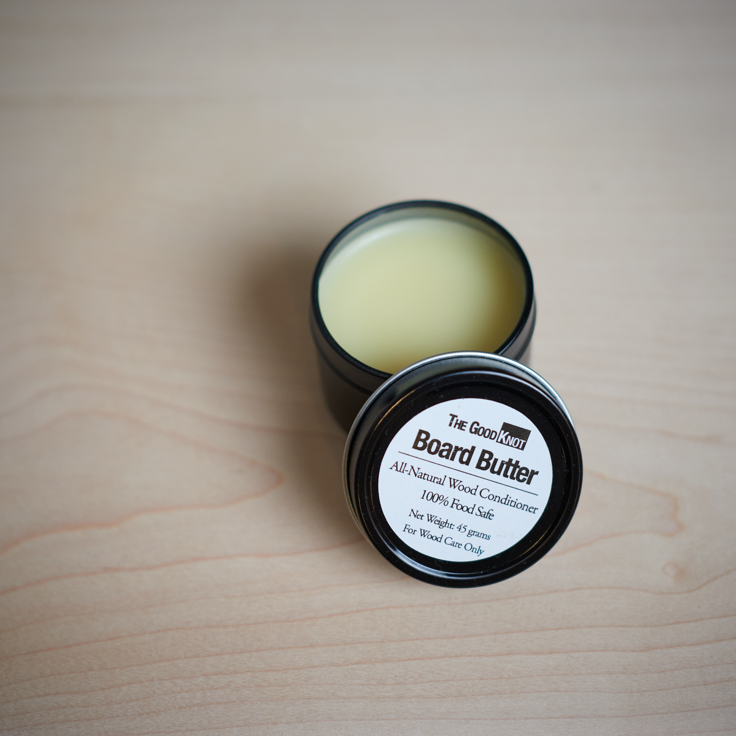 A container of all-natural wood conditioner, Board Butter, with a label that reads 'The Good Knot', is shown on a wooden surface.