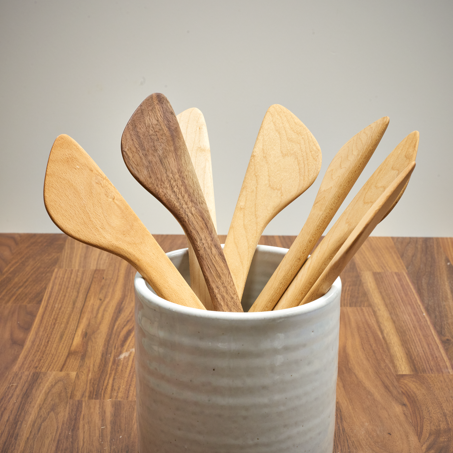 A collection of handcrafted wooden cooking spatulas, displayed upright in a ceramic holder, showcasing different shades and grains of wood.