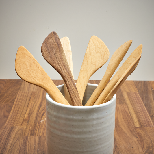 A collection of handcrafted wooden cooking spatulas, displayed upright in a ceramic holder, showcasing different shades and grains of wood.