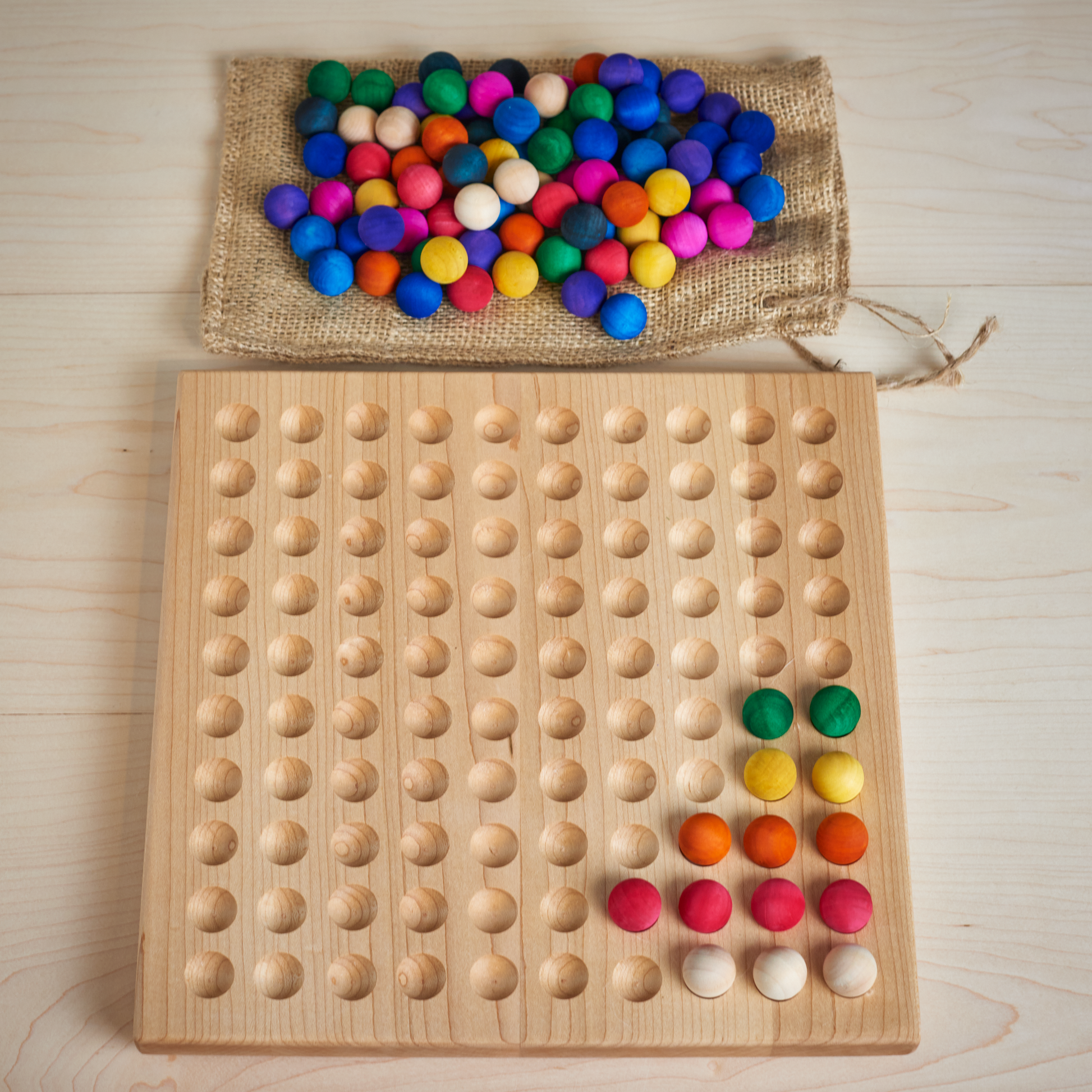 A wooden 100 board with a row of colored wooden balls placed on it, next to a burlap storage bag with more balls.